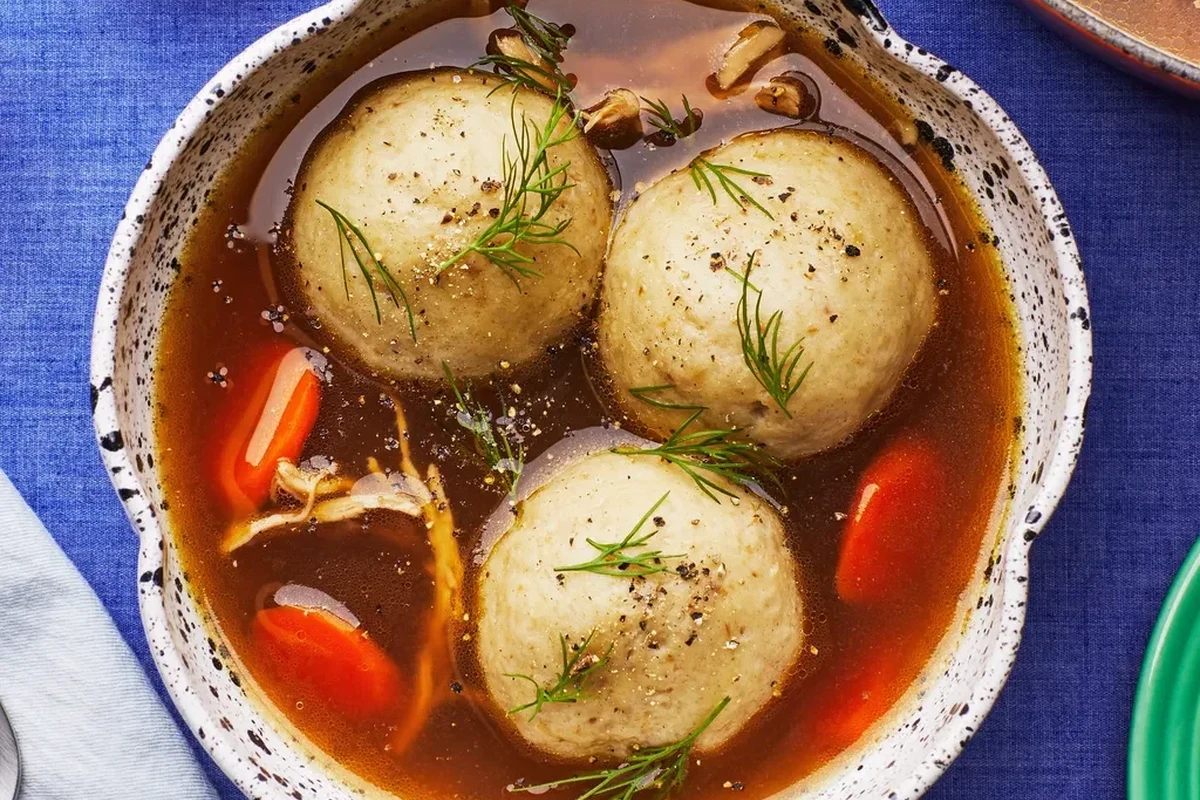 A steaming bowl of clear golden chicken broth with a single, light, fluffy matzo ball and fresh dill garnish.