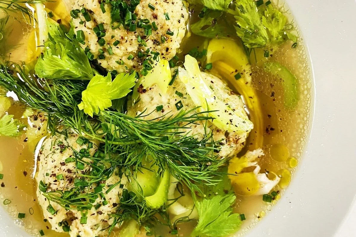 A steaming bowl of clear golden chicken broth with a single, light, fluffy matzo ball and fresh dill garnish.