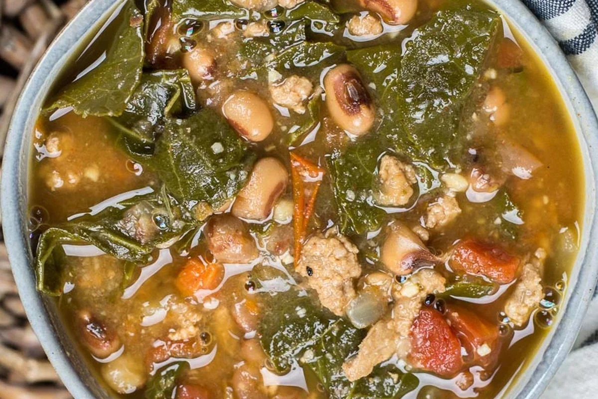 A steaming bowl of black eyed pea and collard green soup with fresh herbs on a rustic wooden table.
