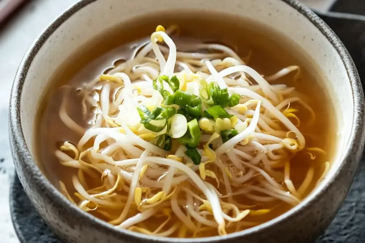A steaming bowl of clear Korean bean sprout soup topped with chopped scallions and chili flakes.