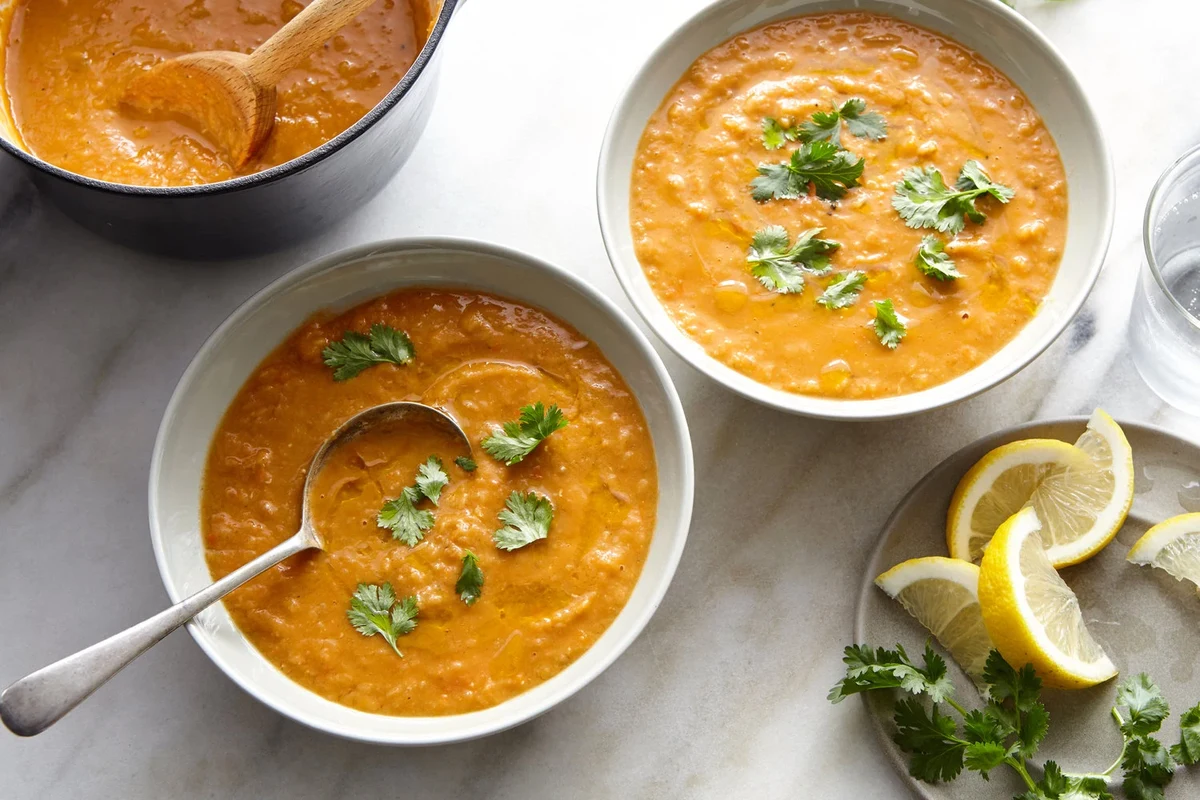 A bowl of creamy, golden red lentil soup garnished with fresh herbs and a slice of lemon on a rustic kitchen table.