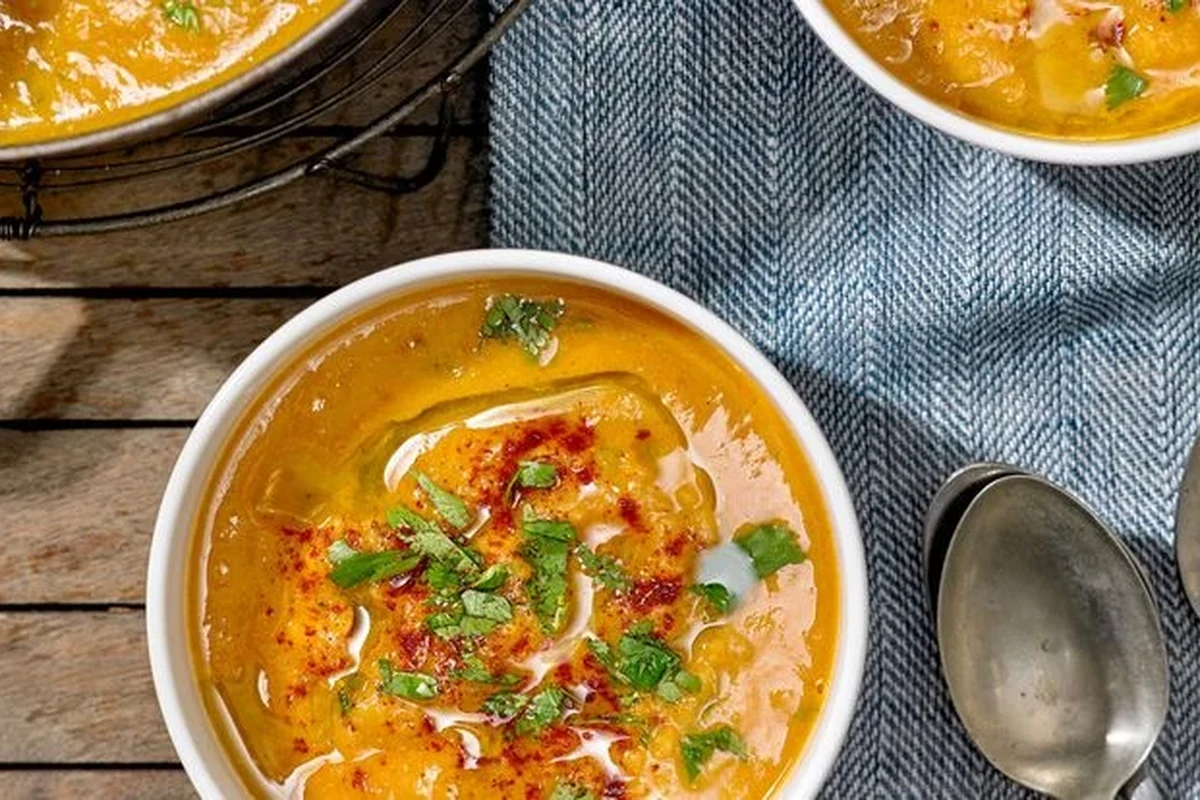 A bowl of creamy, golden red lentil soup garnished with fresh herbs and a slice of lemon on a rustic kitchen table.