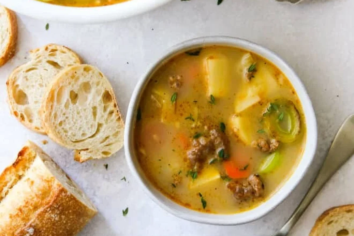 A rustic bowl of potato sausage leek soup topped with fresh herbs on a wooden table.