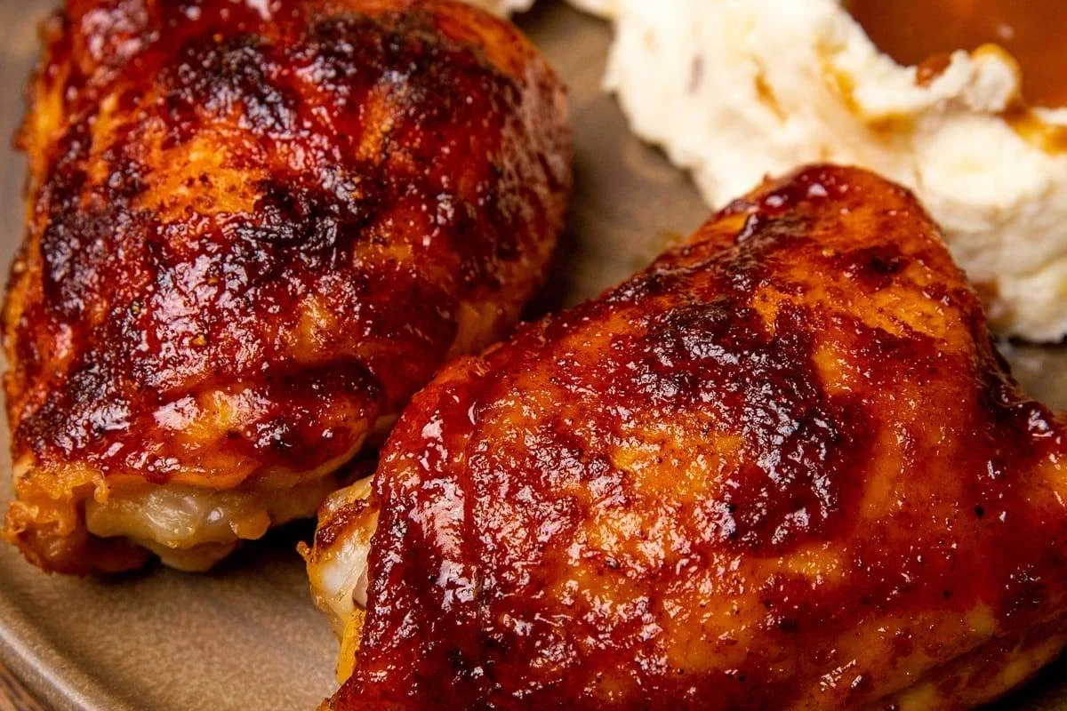 A platter of oven-baked BBQ chicken thighs with a sticky, caramelized glaze on a rustic wooden table.