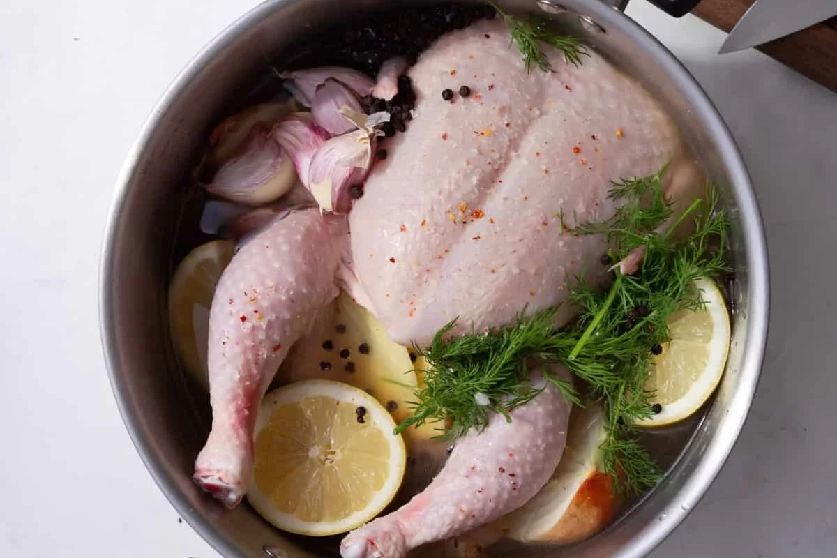 A large pot of water on a stove with a whole chicken simmering alongside fresh aromatic vegetables.