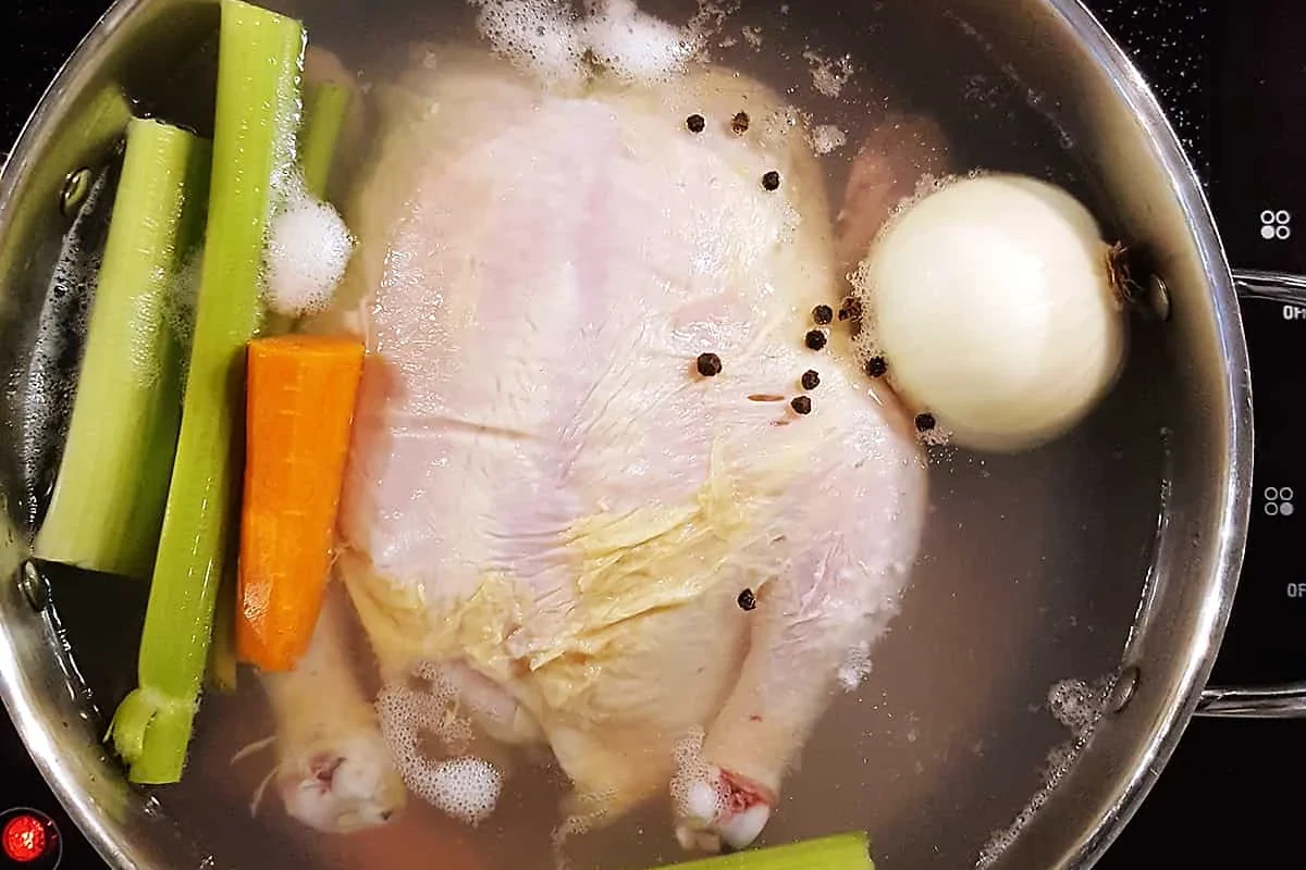 A whole chicken simmering in a large pot with aromatic vegetables like carrots, onions, and celery.