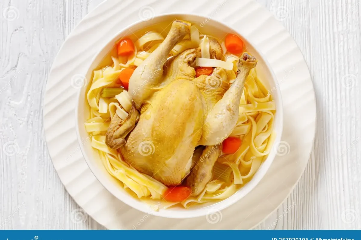 A large pot of golden homemade chicken noodle soup with a whole chicken, fresh herbs, and vegetables on a wooden table.