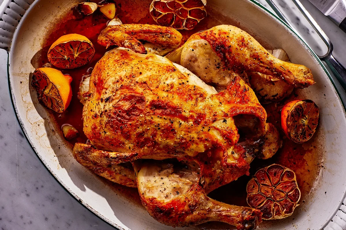 A golden-brown roasted chicken resting on a vertical stainless steel chicken roaster in a kitchen.