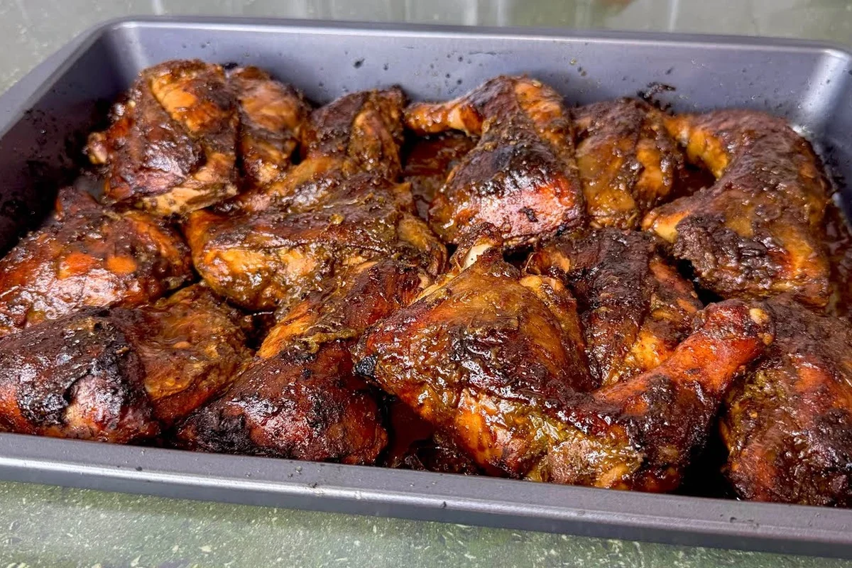 A platter of oven-baked jerk chicken with charred skin, served with fresh thyme and lime wedges.