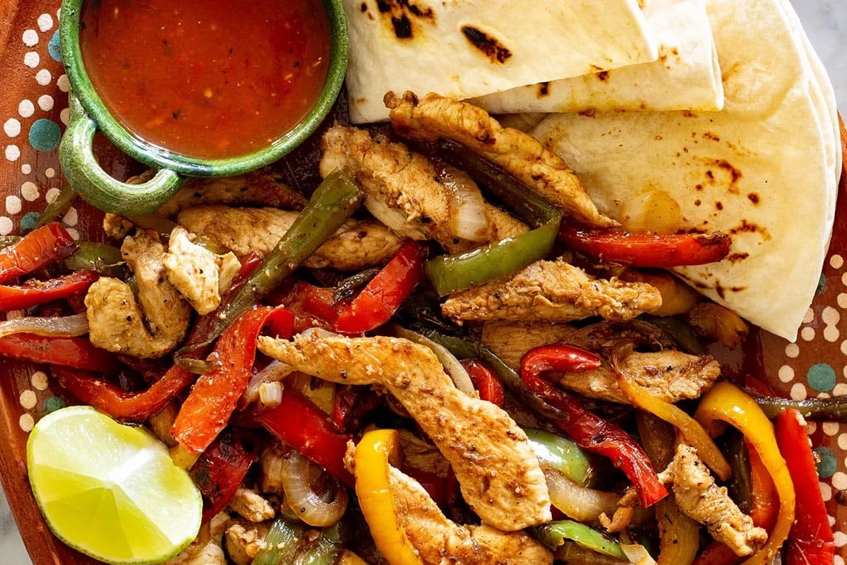 A cast-iron skillet containing sizzling strips of marinated chicken, bell peppers, and onions, served with lime wedges.