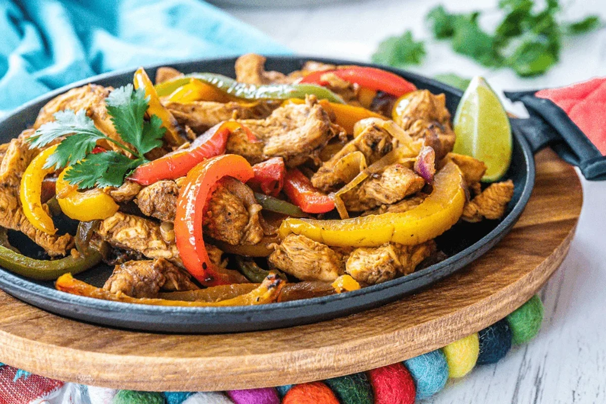 A cast-iron skillet containing sizzling strips of marinated chicken, bell peppers, and onions, served with lime wedges.
