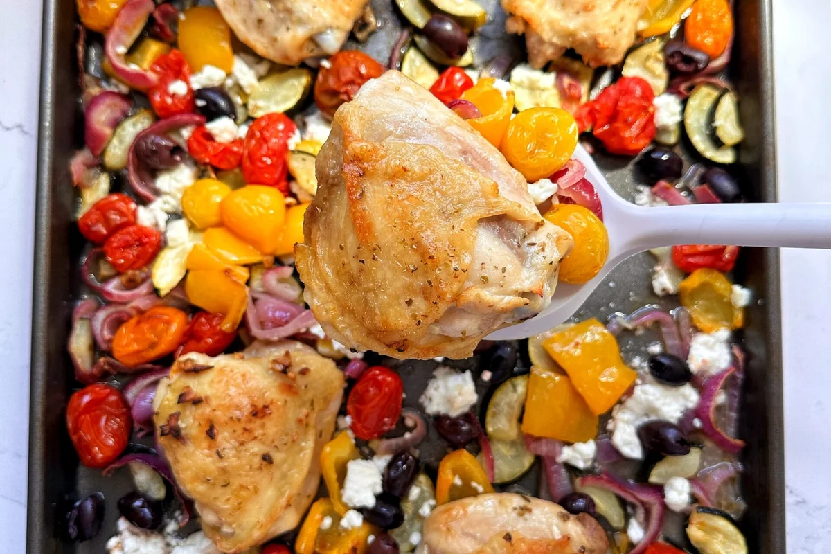 A colorful sheet pan filled with roasted chicken thighs, bell peppers, red onions, and olives, garnished with fresh herbs.