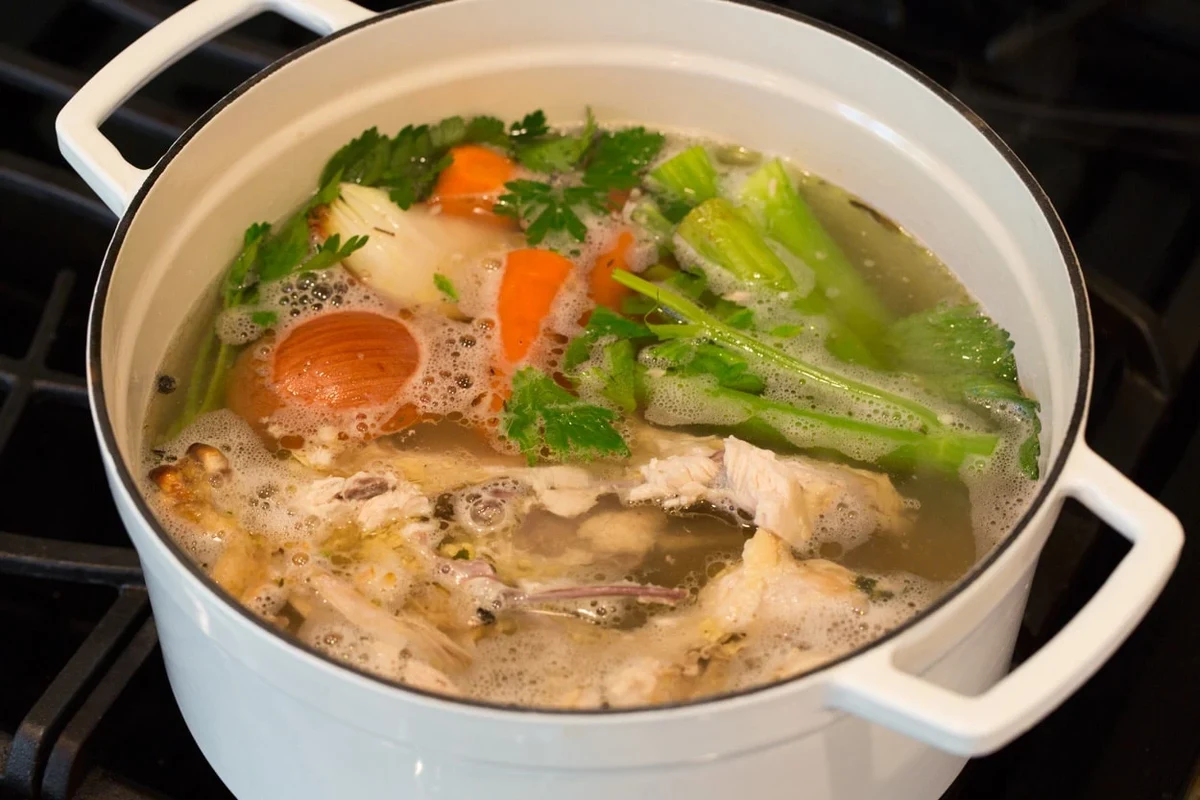 A clear, golden homemade chicken stock in a glass jar surrounded by fresh herbs and aromatics on a wooden kitchen counter.